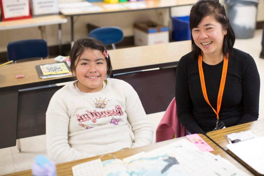 志愿者 tutor and a student sitting at a desk in a classroom side-by-side smiling. 